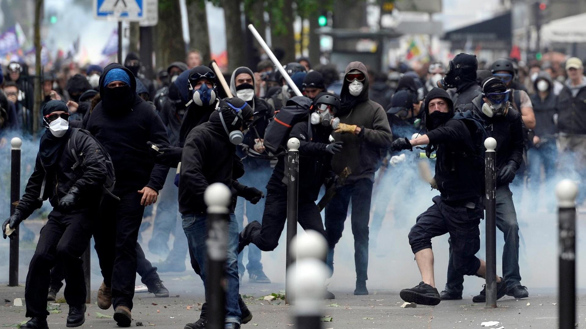 Die teils maskierten schwarzgekleideten Demonstranten werfen Steine und halten weiße Knüppel in den Händen; einige tragen Atemschutz.