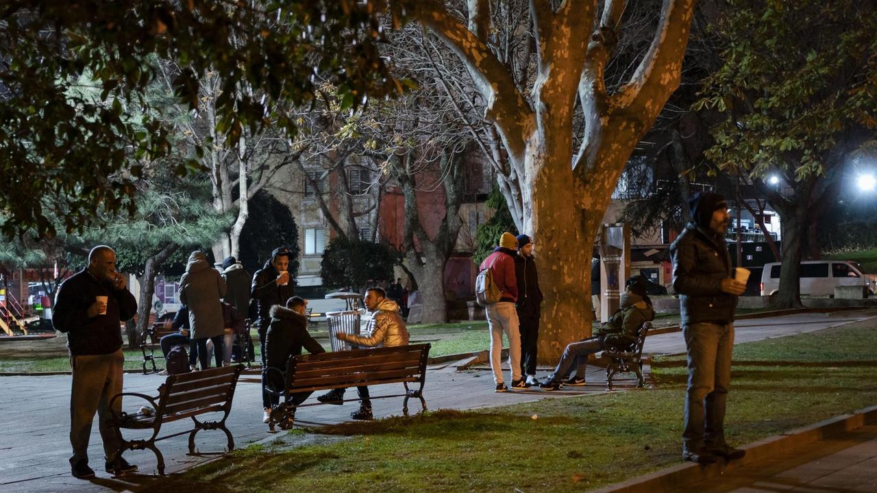 Mehrere Männer im Anorak stehen oder sitzen im Halbdunkel eines Parks und essen und trinken. Mehrere Männer im Anorak stehen oder sitzen im Halbdunkel eines Parks und essen und trinken.