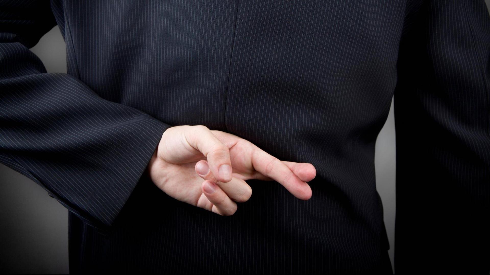 Ein Mann im Anzug hält seine Hand mit gekreuzten Fingern hinter den Rücken