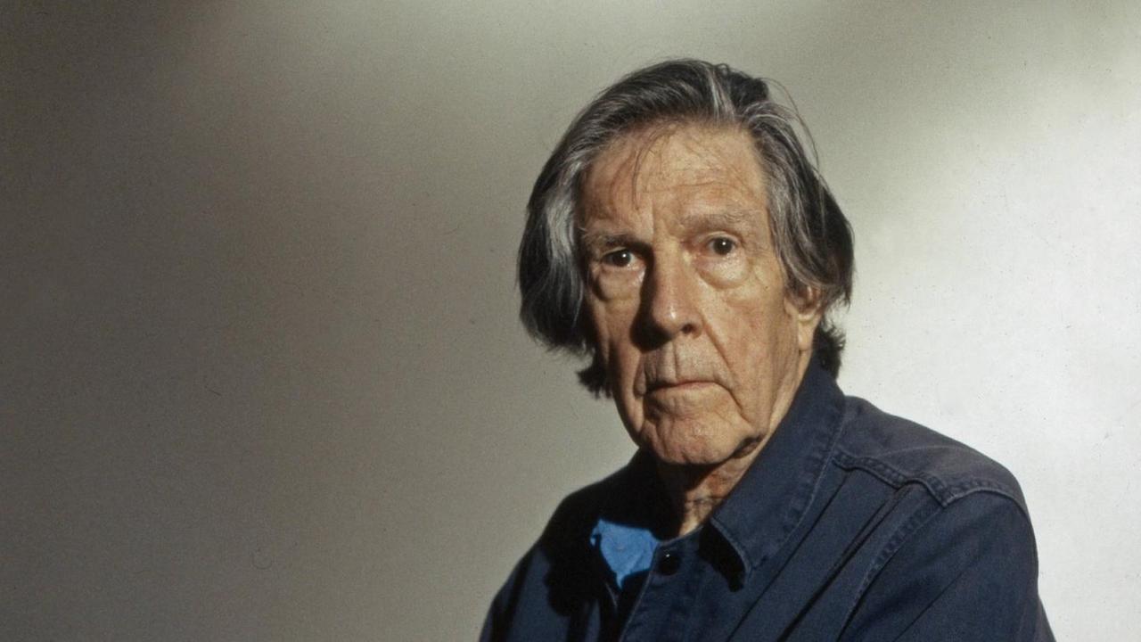 John Cage sitzt seitlich auf einem Stuhl und blickt in die Kamera John Cage sitzt seitlich auf einem Stuhl und blickt in die Kamera