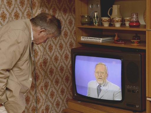 Ein Besucher des DDR-Museums blickt in einen RFT-Fernseher auf dem Eduard Schnitzler zu sehen ist. Dieser steht in einer DDR-typischen Schrankwand in Plattenbauwohnung des Typs WBS 70.