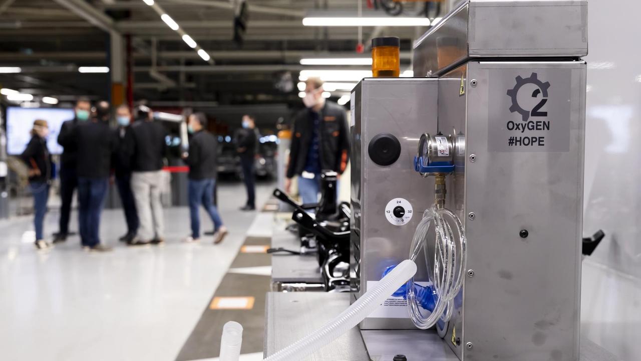 Menschen mit Atemschutzmasken stehen in einer Produktionshalle. Im Vordergrund steht ein großer Metall-Apparat mit Schlauch auf dem "Oxygen" und "Hope" steht. Menschen mit Atemschutzmasken stehen in einer Produktionshalle. Im Vordergrund steht ein großer Metall-Apparat mit Schlauch auf dem "Oxygen" und "Hope" steht.