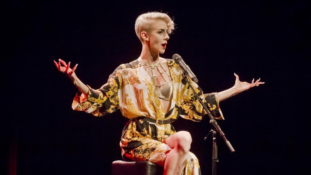 Lisa Eckhart, mit kurzen blonden Haaren in einem schwarz-gelbem Gewand sitzt auf einer Bühne und spricht in ein Mikrofon.
