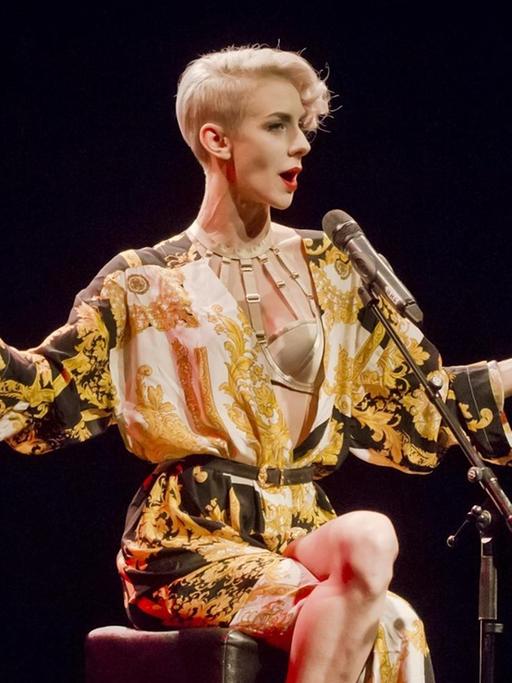 Lisa Eckhart, mit kurzen blonden Haaren in einem schwarz-gelbem Gewand sitzt auf einer Bühne und spricht in ein Mikrofon.