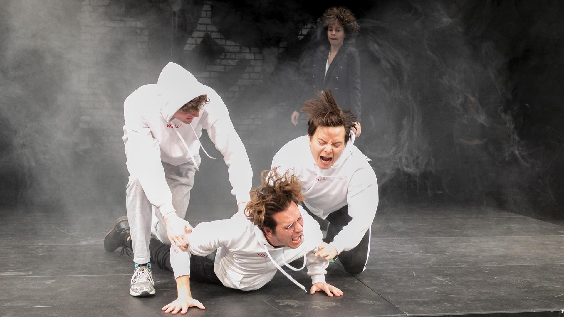 Szenenfoto aus "Die Welle" am Grips Theater in Berlin: Im Vordergrund drei junge Männer in weißen Kapuzenpullovern, einer liegt auf dem Boden und die anderen helfen ihm entweder nach oben oder drücken ihn nach unten. Szenenfoto aus "Die Welle" am Grips Theater in Berlin: Im Vordergrund drei junge Männer in weißen Kapuzenpullovern, einer liegt auf dem Boden und die anderen helfen ihm entweder nach oben oder drücken ihn nach unten.
