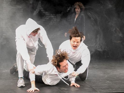 Szenenfoto aus "Die Welle" am Grips Theater in Berlin:  Im Vordergrund drei junge Männer in weißen Kapuzenpullovern, einer liegt auf dem Boden und die anderen helfen ihm entweder nach oben oder drücken ihn nach unten. 