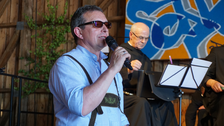 Hubert steht auf der Bühne und spricht ins Mikrofon. Hubert steht auf der Bühne und spricht ins Mikrofon.