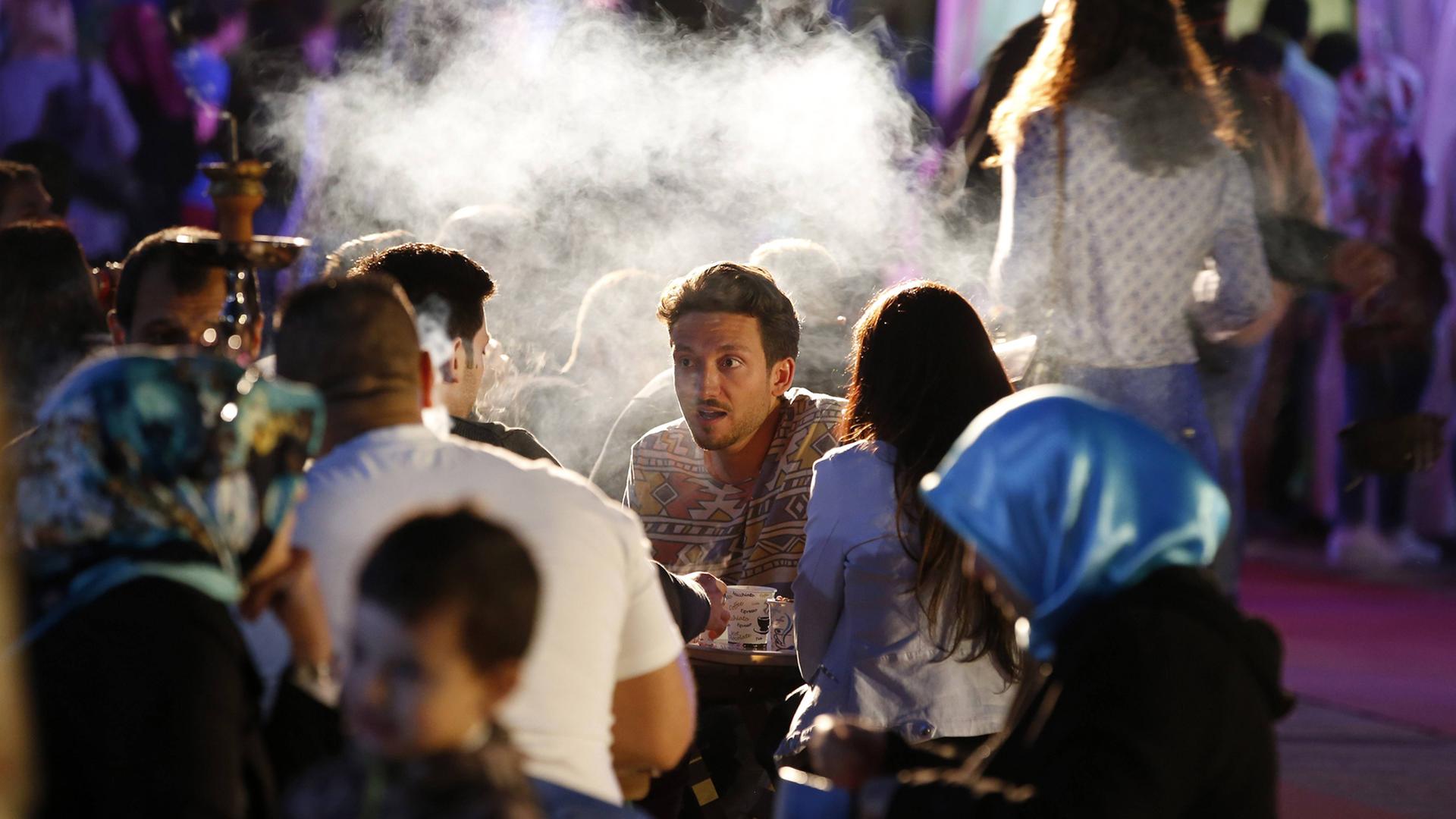 Menschen sitzen an langen Tischen, der Fokus liegt auf einem jungen Mann, der mit anderen spricht, Dampf aus einer Shisha steigt auf. Menschen sitzen an langen Tischen, der Fokus liegt auf einem jungen Mann, der mit anderen spricht, Dampf aus einer Shisha steigt auf.