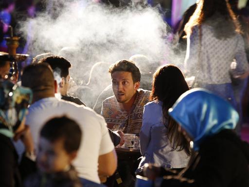Menschen sitzen an langen Tischen, der Fokus liegt auf einem jungen Mann, der mit anderen spricht, Dampf aus einer Shisha steigt auf.