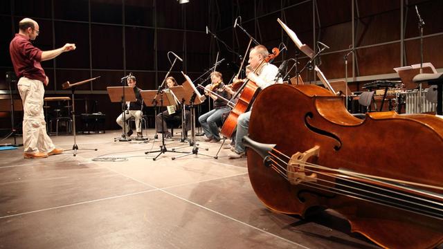 Fünf Musik stizen mit Instrumenten vor einem Dirigenten auf einer Bühne. Rechts von Ihnen liegt ein Kontrabass auf dem Boden.