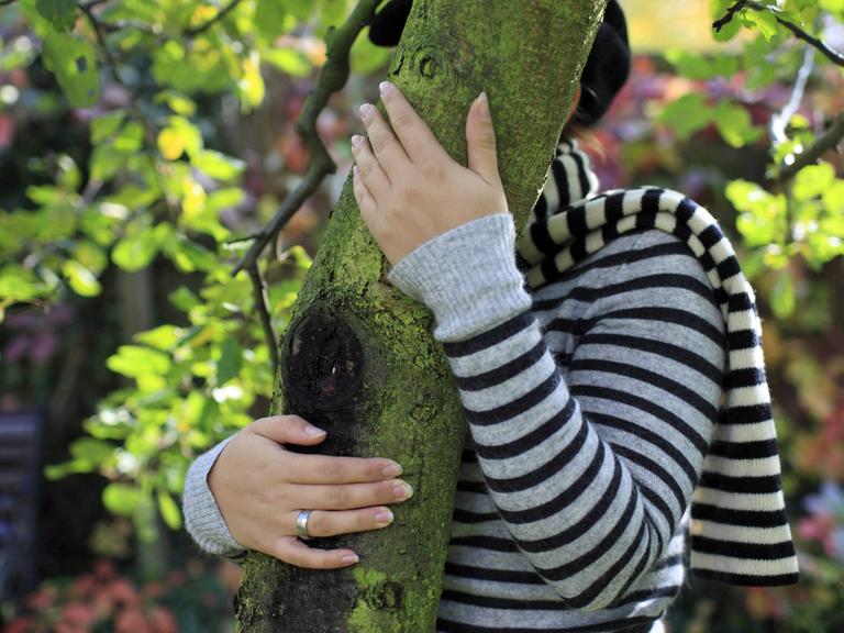 Eine junge Frau versteckt sich hinter einem Baumstamm.