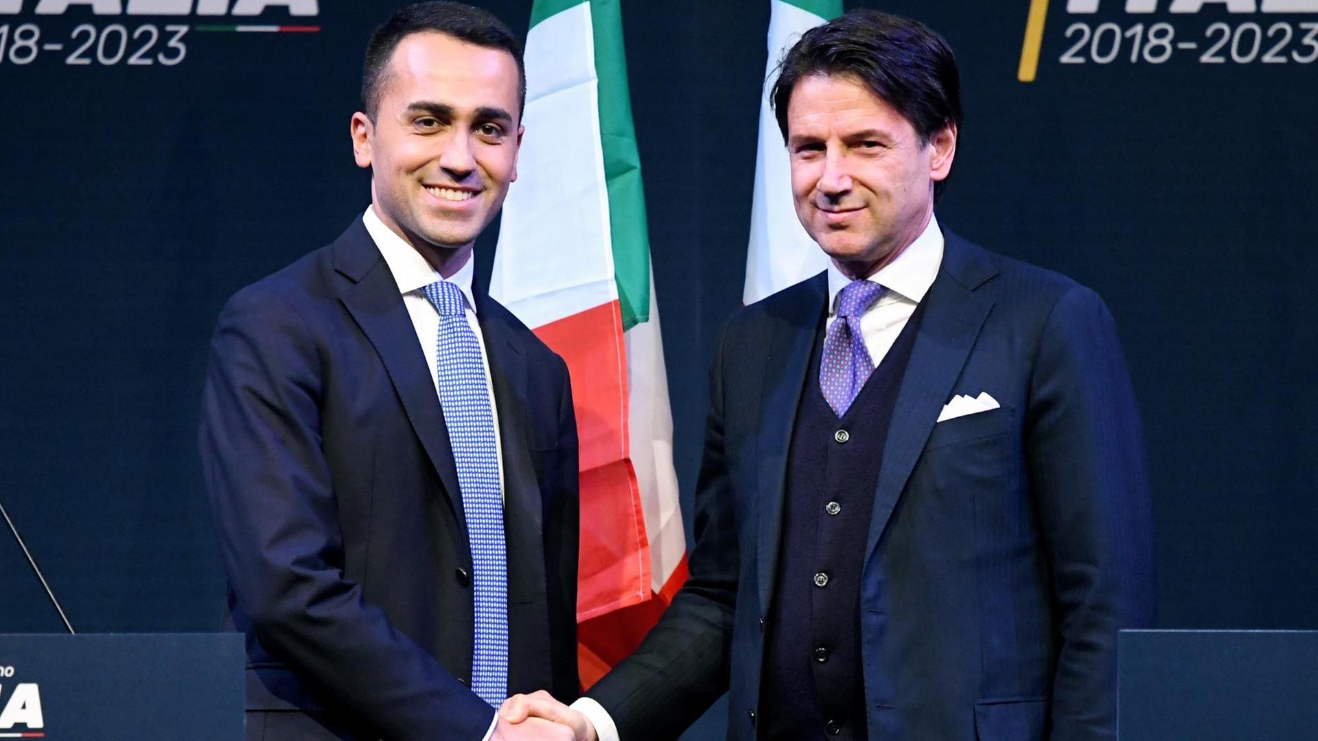 Die beiden stehen vor einer schwarzen Wand mit zwei italienischen Fahnen und schütteln sich lächelnd für die Fotografen die Hände. Die beiden stehen vor einer schwarzen Wand mit zwei italienischen Fahnen und schütteln sich lächelnd für die Fotografen die Hände.