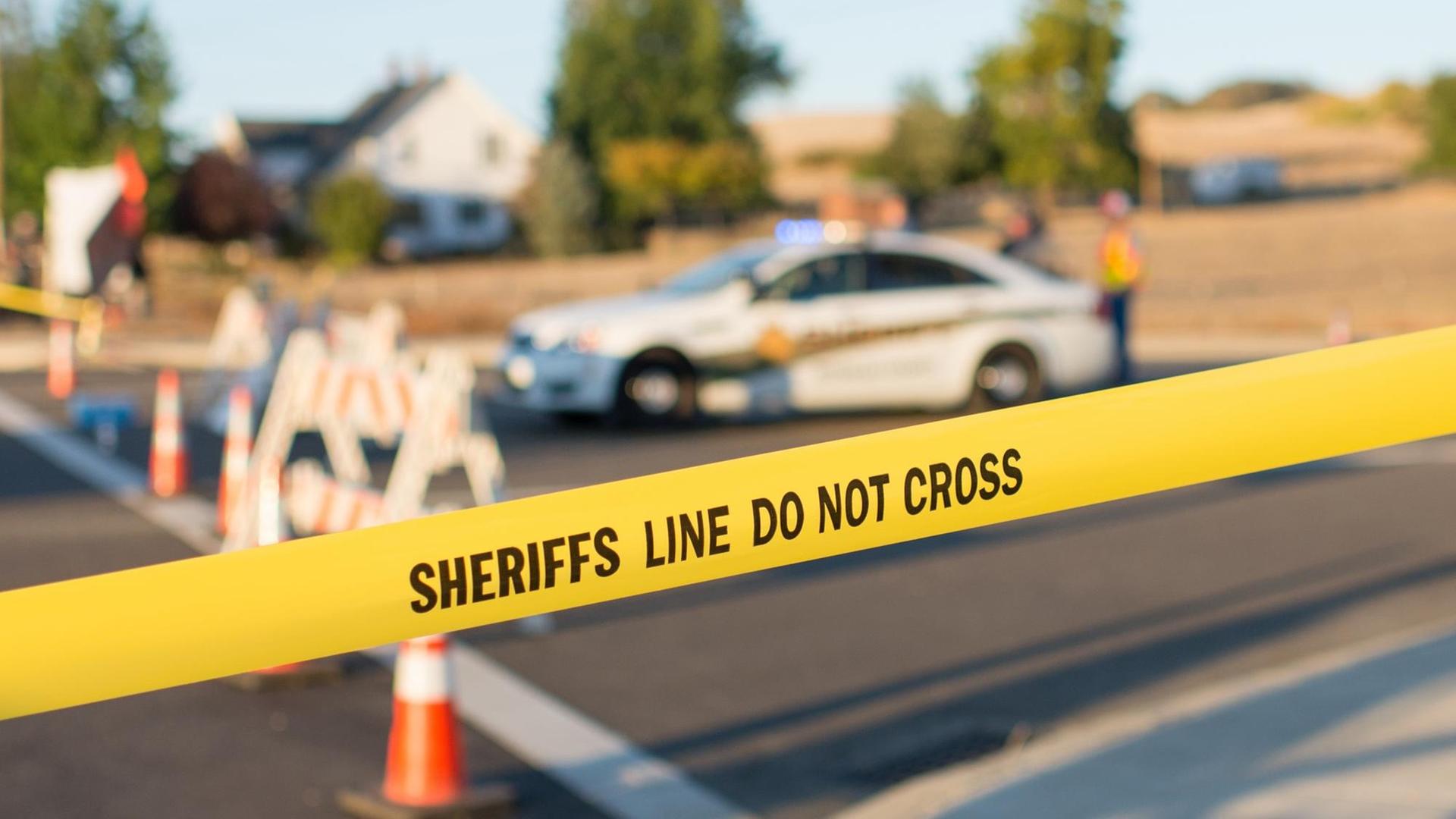 Hinter der Absperrung mit der Aufschrift "Sheriffs Line do not cross" ist ein Polizeiauto zu sehen. Hinter der Absperrung mit der Aufschrift "Sheriffs Line do not cross" ist ein Polizeiauto zu sehen.