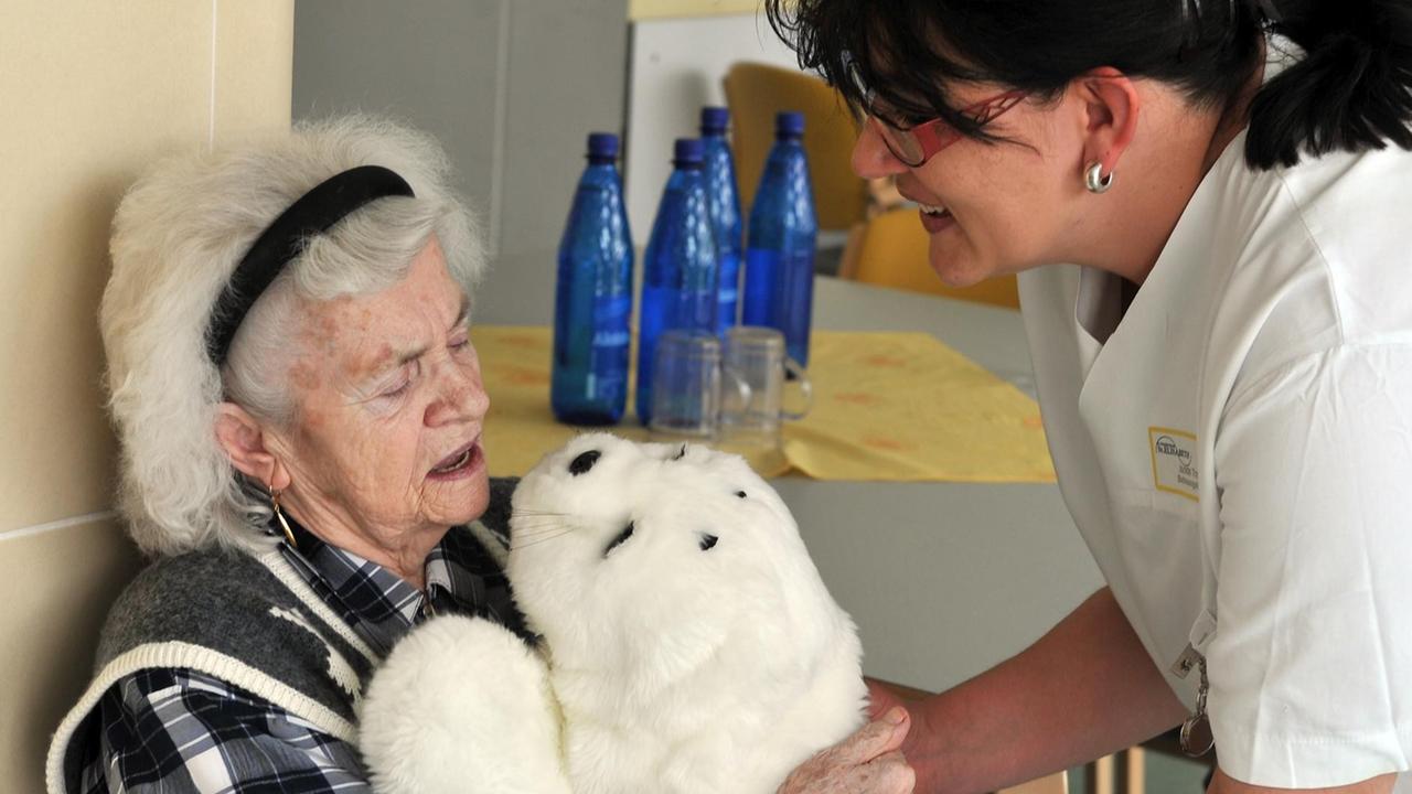 Eine Pflegekraft reicht der dementen Eleonore Mayer einen Roboter in einem Robbenfell, der sich bewegt und Geräusche macht Eine Pflegekraft reicht der dementen Eleonore Mayer einen Roboter in einem Robbenfell, der sich bewegt und Geräusche macht