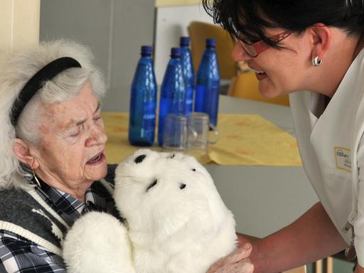 Eine Pflegekraft reicht der dementen Eleonore Mayer einen Roboter in einem Robbenfell.