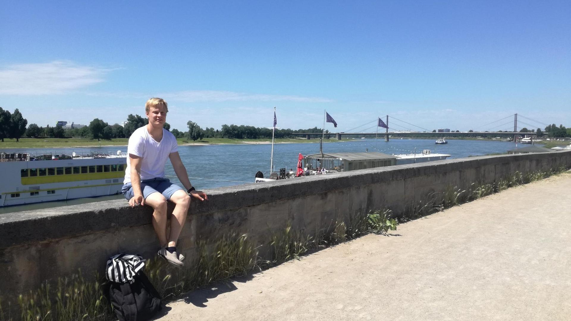 Ein junger Mann sitzt auf einer Mauer am Fluss Rhein. Hinter ihm sieht man Schiffe und eine Brücke. Ein junger Mann sitzt auf einer Mauer am Fluss Rhein. Hinter ihm sieht man Schiffe und eine Brücke.