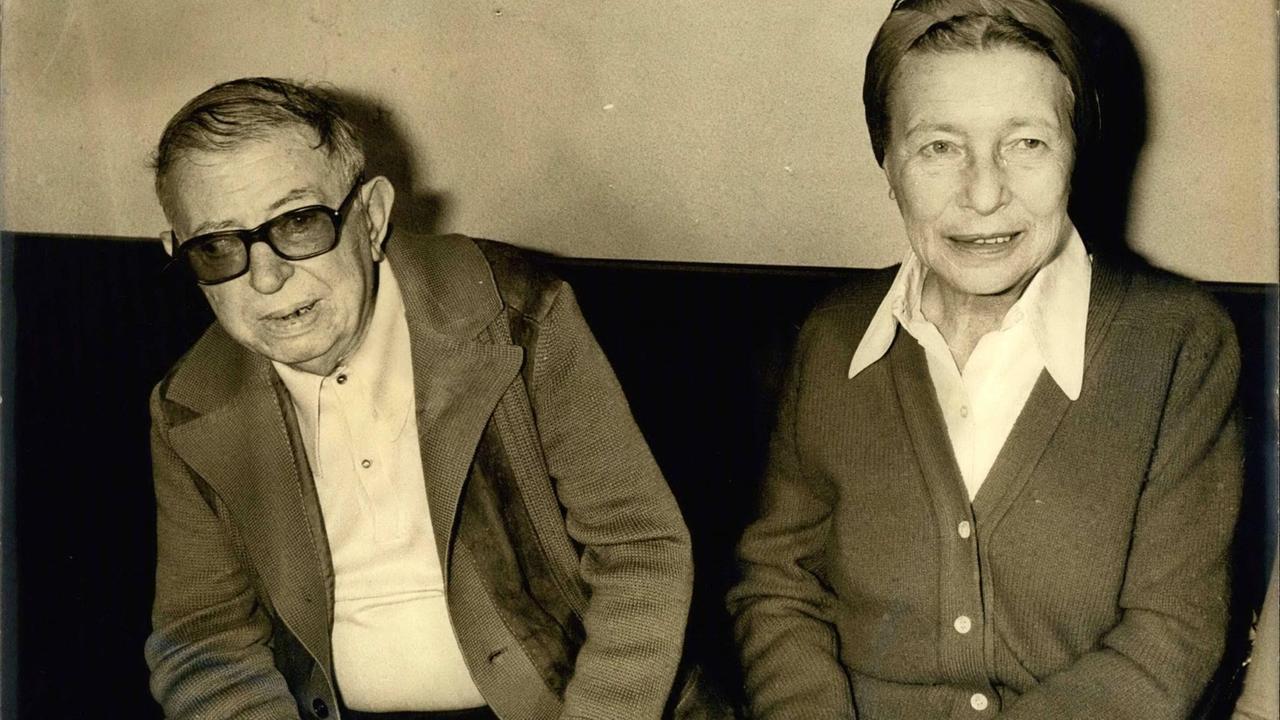 Jean-Paul Sartre und Simone de Beauvoir sitzen nebeinander.De Beauvoir schaut lächelnd zur Seite, Sartre schaut direkt in die Kamera. Jean-Paul Sartre und Simone de Beauvoir sitzen nebeinander.De Beauvoir schaut lächelnd zur Seite, Sartre schaut direkt in die Kamera.
