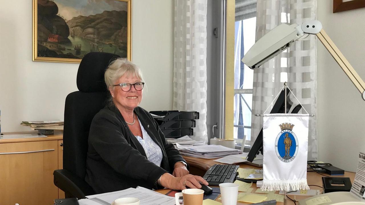 Eine Frau mit blondem hochgestecktem Haar und Brille sitzt lächelnd an ihrem Schreibtisch vor ihrem PC. Eine Frau mit blondem hochgestecktem Haar und Brille sitzt lächelnd an ihrem Schreibtisch vor ihrem PC.