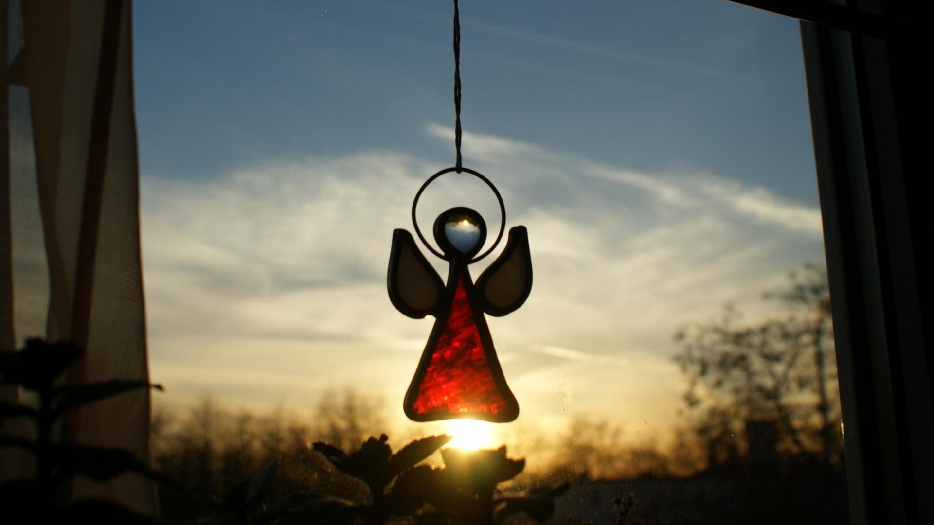 Eine Engelsfigur aus Glas hängt am Fenster, dahinter ein Sonnenuntergang. Eine Engelsfigur aus Glas hängt am Fenster, dahinter ein Sonnenuntergang.