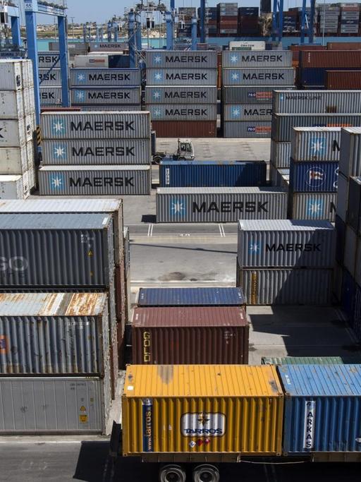 Das Bild zeigt viele übereinandergestapelte Containertürme, die meisten mit der Aufschrift "Maersk". Im Hintergrund Kräne und ein Hafenbecken.