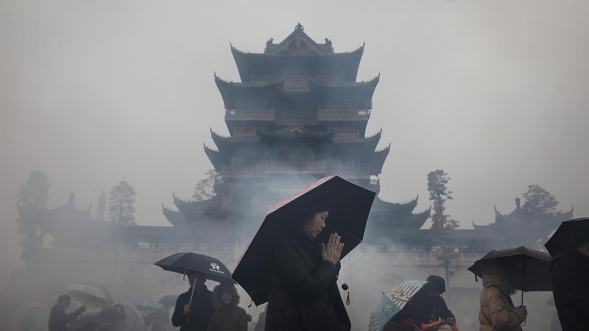 Eine Frau mit einem Regenschirm betet vor der Silhouette eines buddhistischen Tempels.