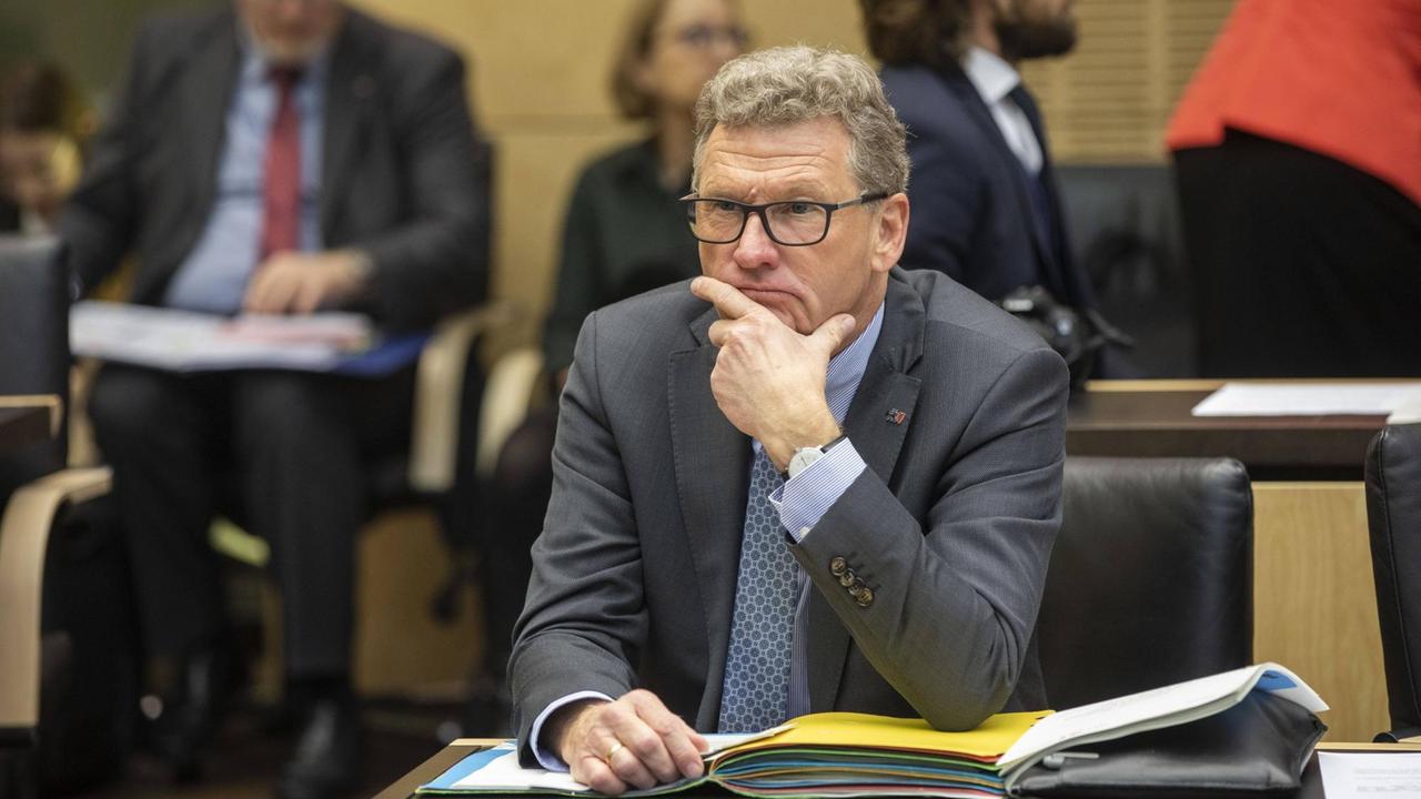 Ein Mann mit Brille und kurzen hellen Haaren in einem Sitzungssaal und hat das Kinn auf die Hand gestützt. Vor ihm auf dem Tisch liegen Aktenordner. Ein Mann mit Brille und kurzen hellen Haaren in einem Sitzungssaal und hat das Kinn auf die Hand gestützt. Vor ihm auf dem Tisch liegen Aktenordner.