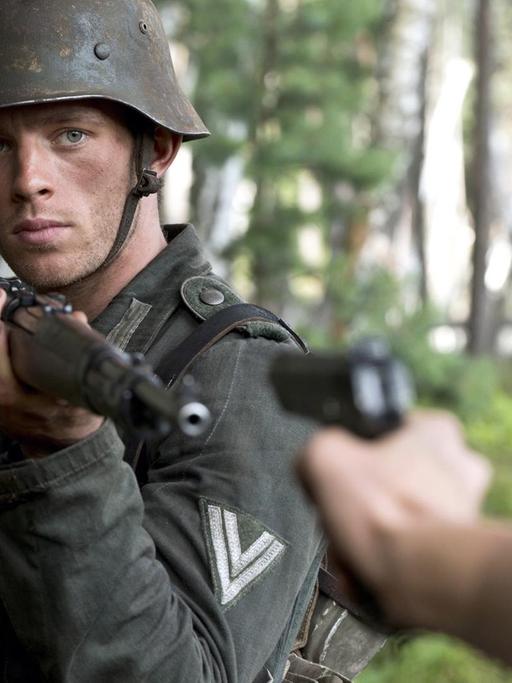 Ein Wehrmachtssoldat richtet das Gewehr auf eine Person, die ihn mit einer Pistole bedroht.