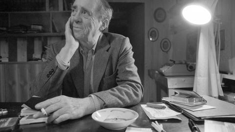 Eine historische Aufnahme vom deutschen Schriftsteller Heinrich Böll. Er sitzt rauchend und nachdenklich am Schreibtisch in seiner Wohnung und lächelt verschmitzt.