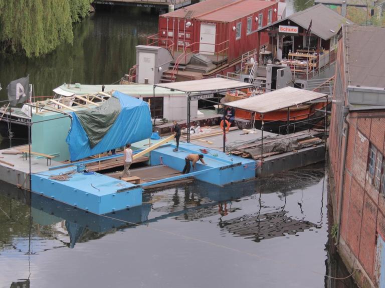 Blaue Pontons schwimmen im Wasser, drauf ein Holzdachaufbau: Die schwimmende Bibliothek im Aufbau am Veringkanal im Hamburger Stadtteil Wilhelmsburg
