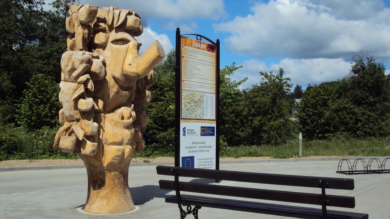 Die aus einem Holzstamm geschnitzte übermannshohe Figur eines Dämons mit erhobenem Rüssel steht an einer Bushaltestelle zwischen einer Sitzbank und einer Informationstafel.