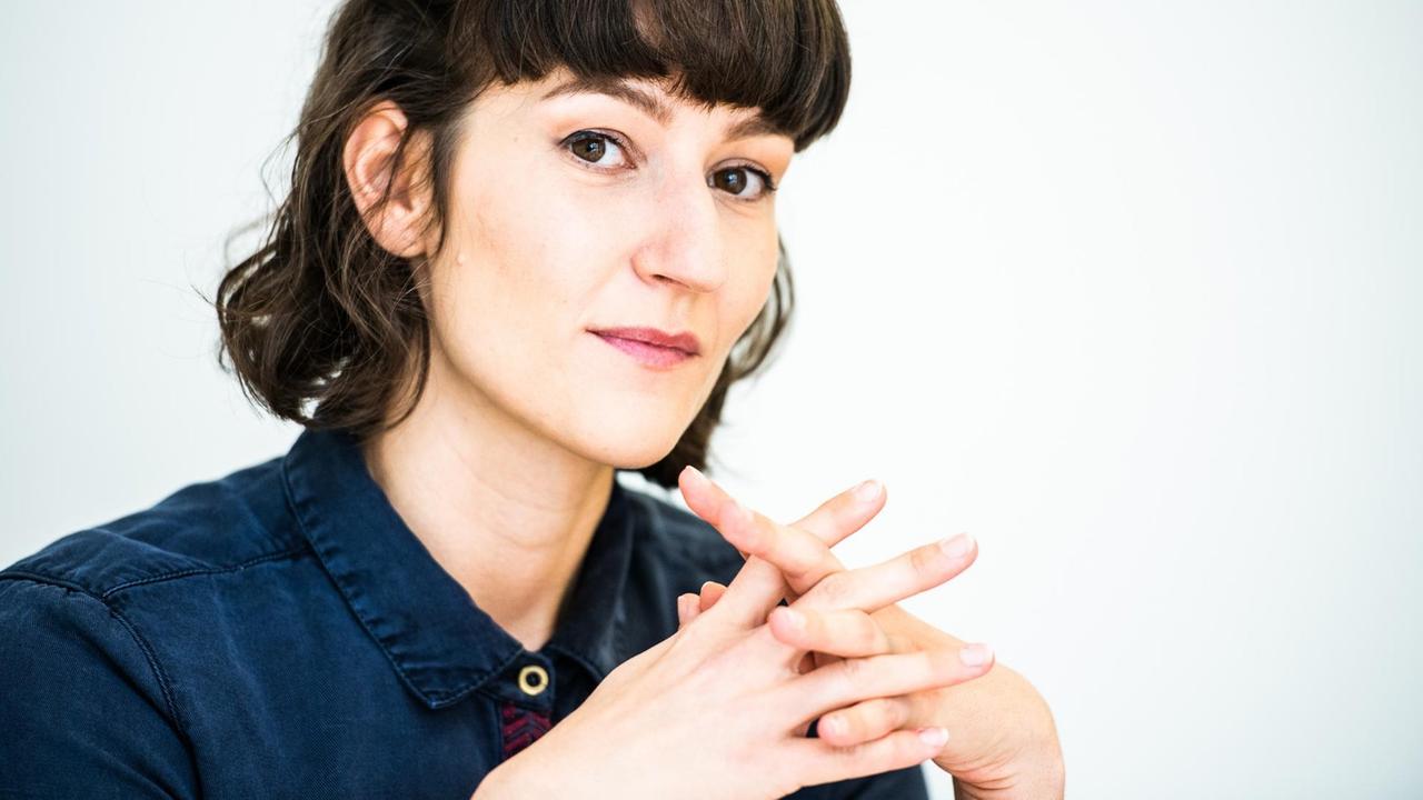 Lena Papasabbas, mit halblangem dunklem Haar, in einem dunkelblauen Hemd, hat die Finger beider Hände ineinander verschränkt und schaut freundlich in die Kamera.