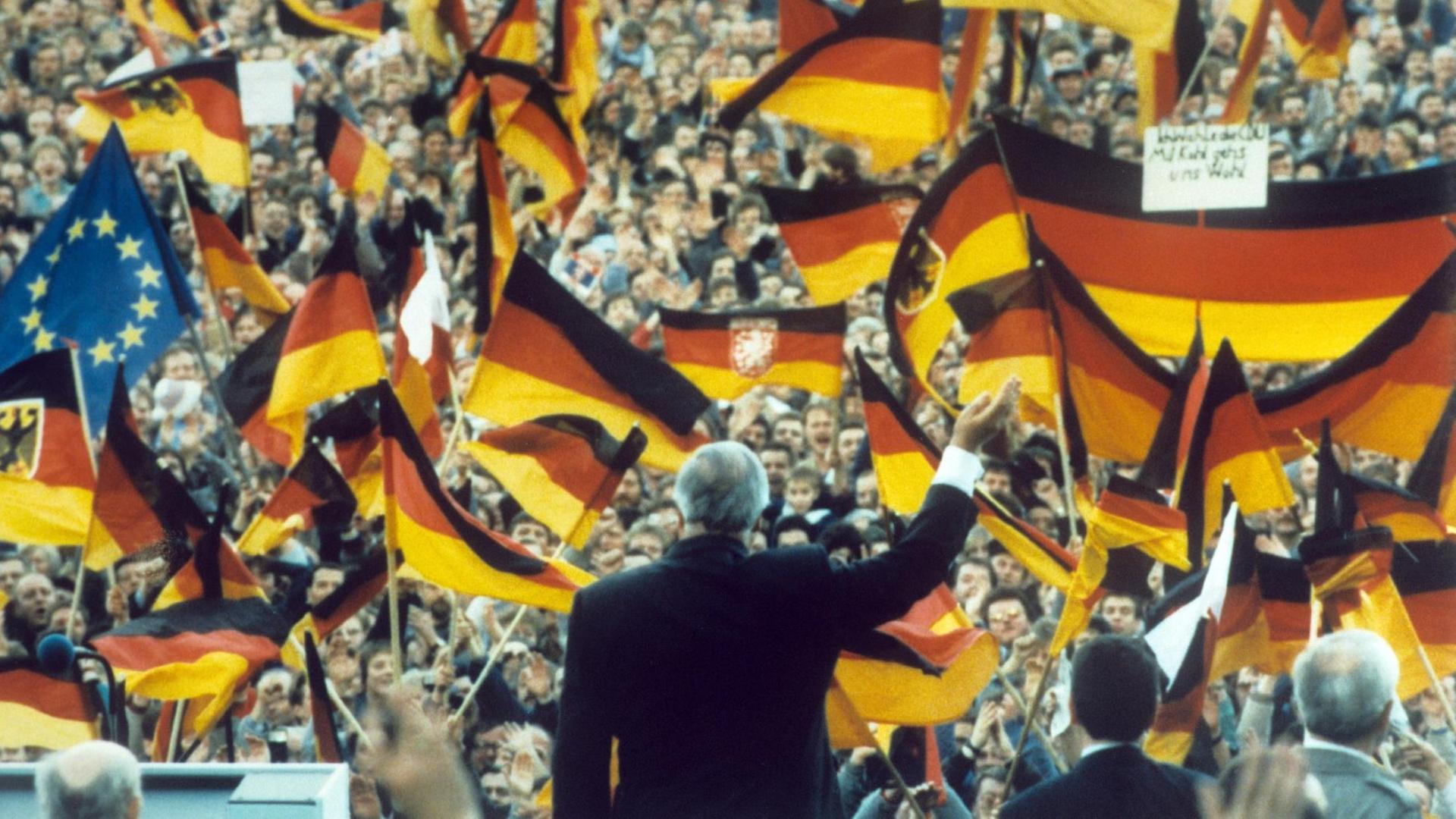 Helmut Kohl grüßt in die Menge. Viele Menschen schwenken Deutschland-Fahnen, auch eine Europa-Fahne ist dabei.