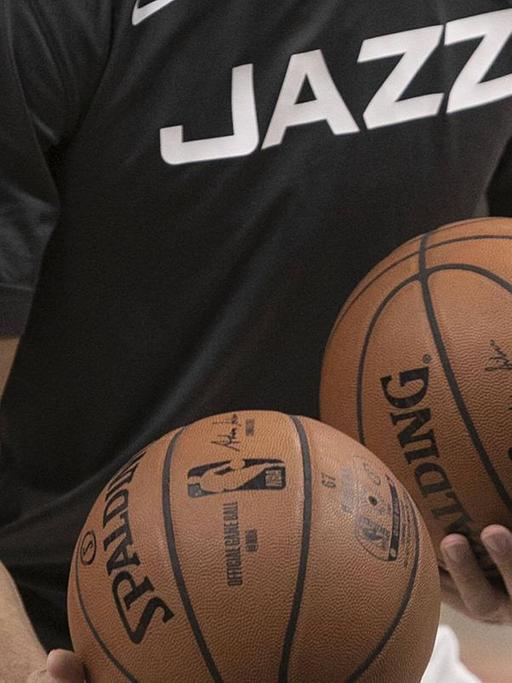 Auf dem Bild ist der Rumpf eines Mannes zu sehen, der ein schwarzes T-Shirt mit der weißen Aufschrift "Jazz" trägt und zwei Basketbälle in den Händen hält.