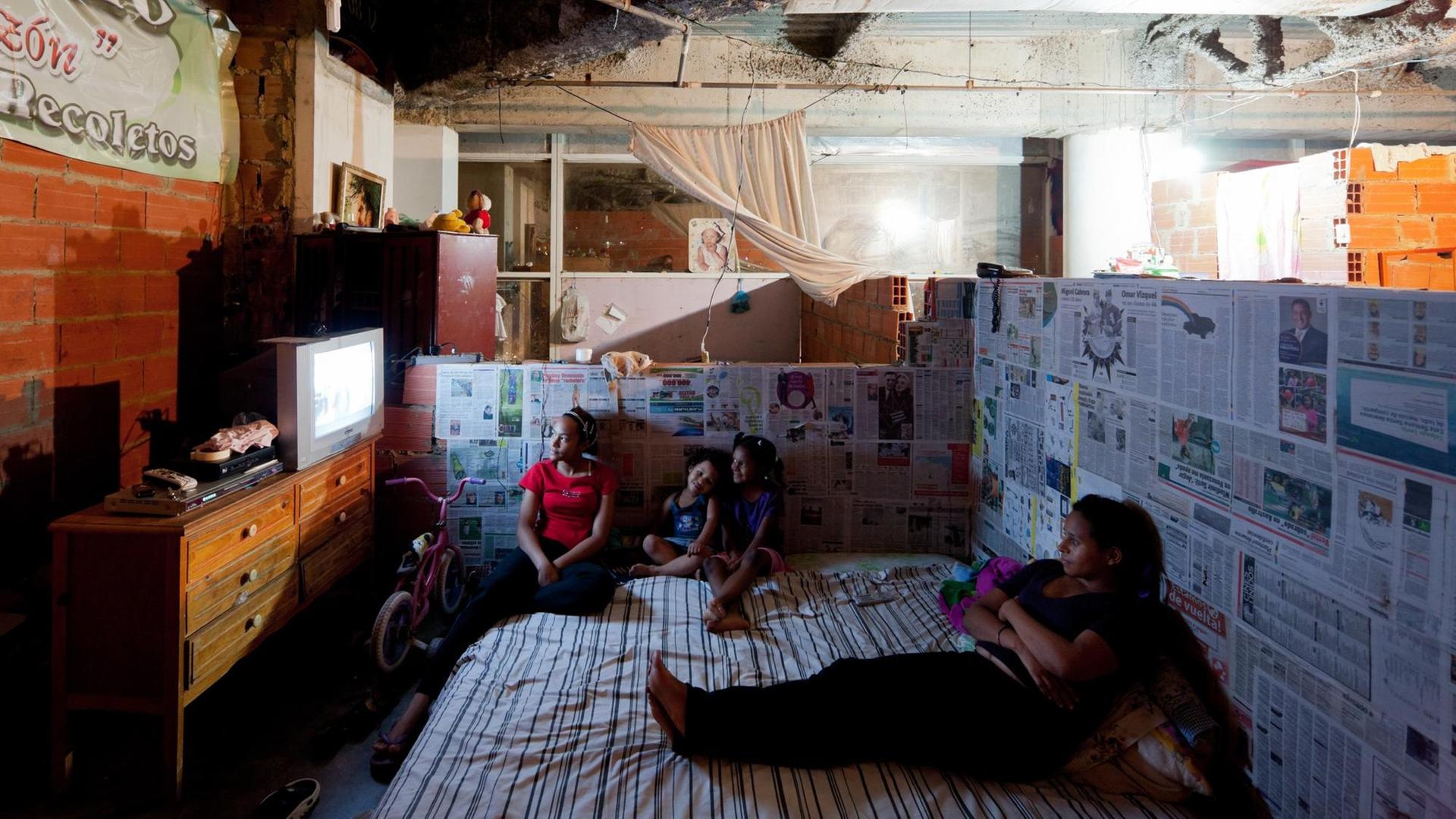Four people sit on mattresses on the floor in a shell and watch television