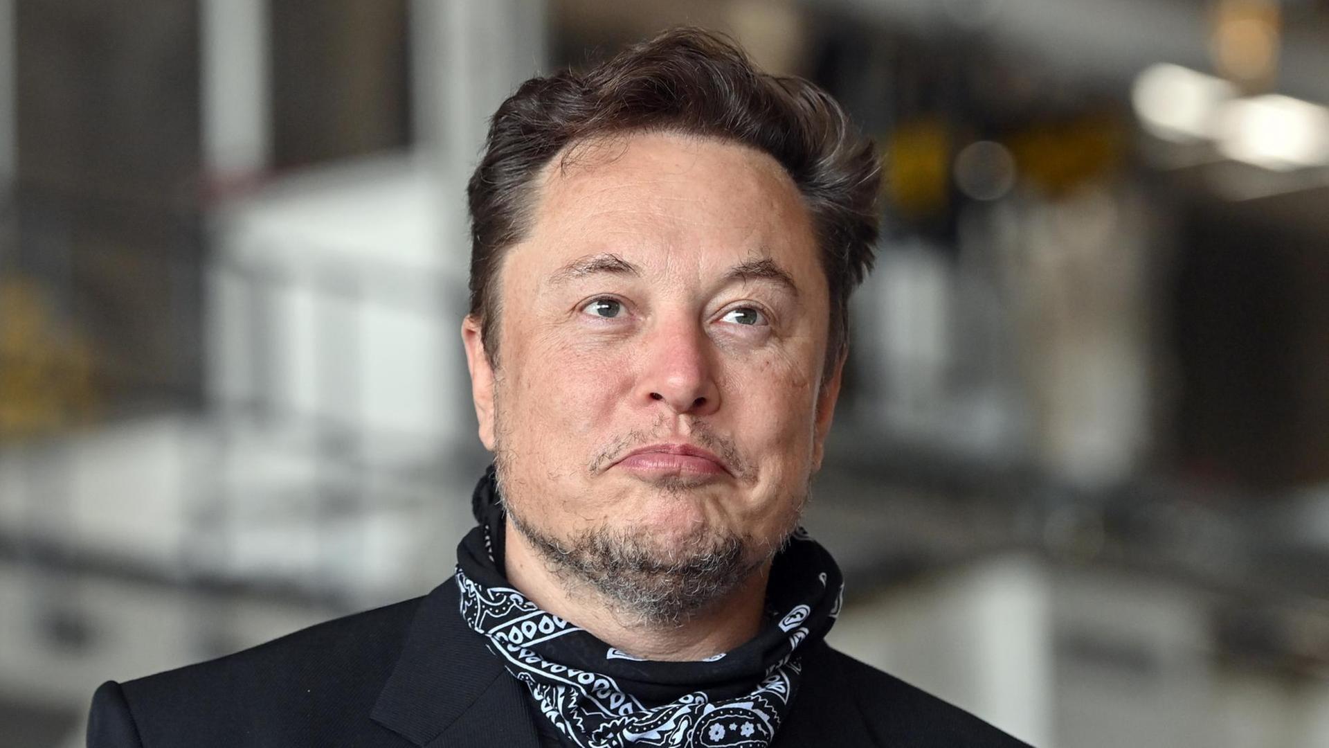 Zu sehen ist Elon Musk mit Drei-Tage-Bart. Er steht in einer Fabrikhalle udn trägt einen schwarzen Anzug und ein Nickituch. Zu sehen ist Elon Musk mit Drei-Tage-Bart. Er steht in einer Fabrikhalle udn trägt einen schwarzen Anzug und ein Nickituch.