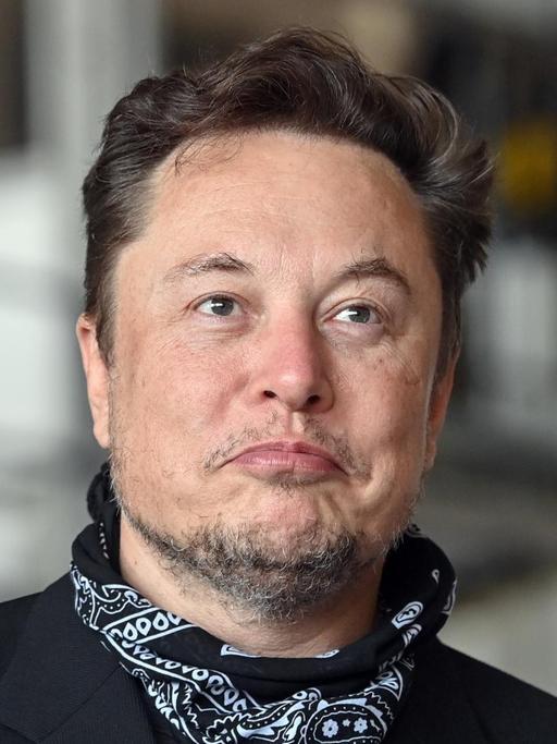 Zu sehen ist Elon Musk mit Drei-Tage-Bart. Er steht in einer Fabrikhalle udn trägt einen schwarzen Anzug und ein Nickituch.