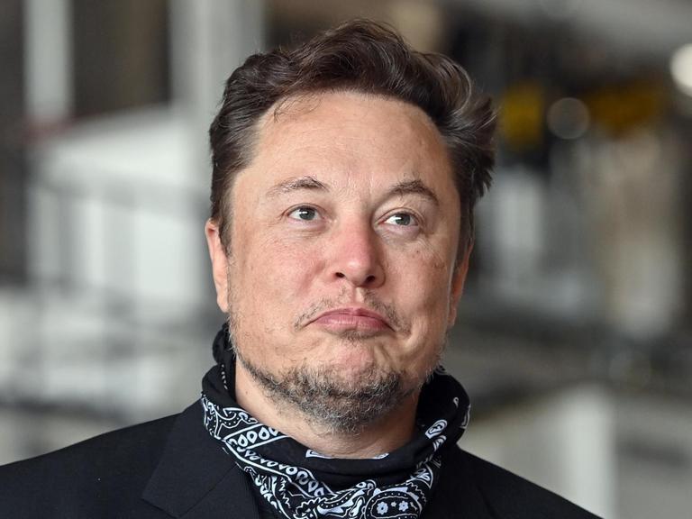 Zu sehen ist Elon Musk mit Drei-Tage-Bart. Er steht in einer Fabrikhalle udn trägt einen schwarzen Anzug und ein Nickituch.