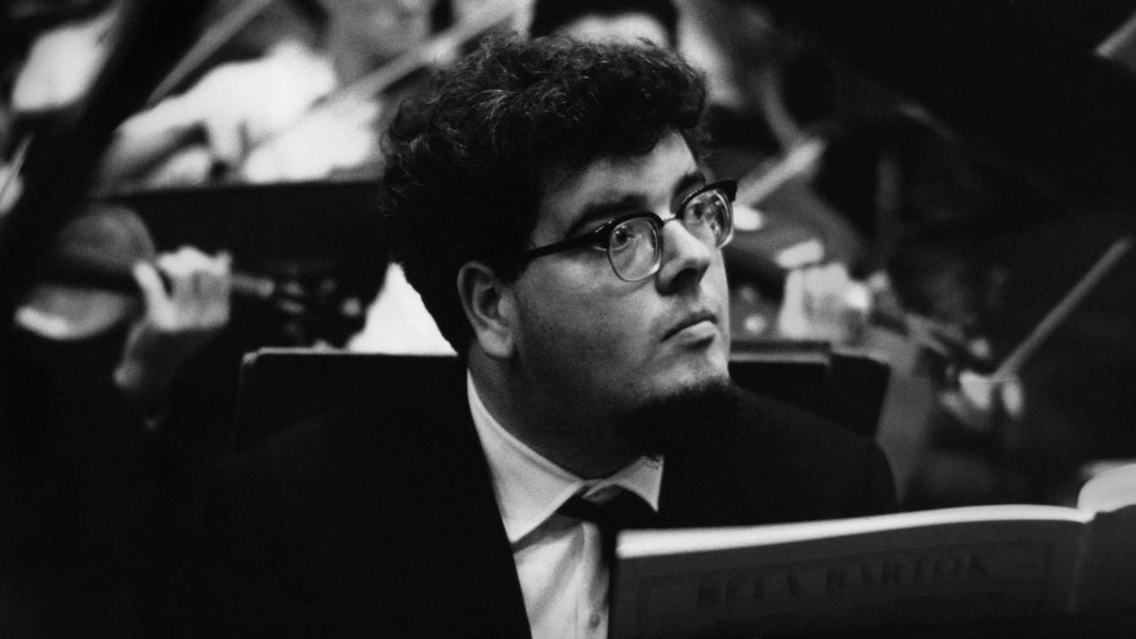 John Ogdon mit Brille und dunklen, kurzen Locken an einem Flügel sitzend, dahinter sind die Geiger eines Orchesters zu erkennen. John Ogdon mit Brille und dunklen, kurzen Locken an einem Flügel sitzend, dahinter sind die Geiger eines Orchesters zu erkennen.