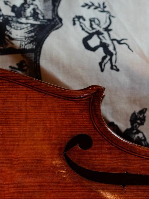 Detail einer barocken Geige, die auf einem mit Putten bedrucktem Tuch liegt.