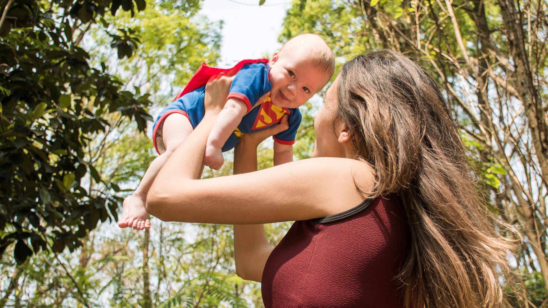 Eine Frau hebt ein Baby in einem Superman-Kostüm in die Luft. Eine Frau hebt ein Baby in einem Superman-Kostüm in die Luft.