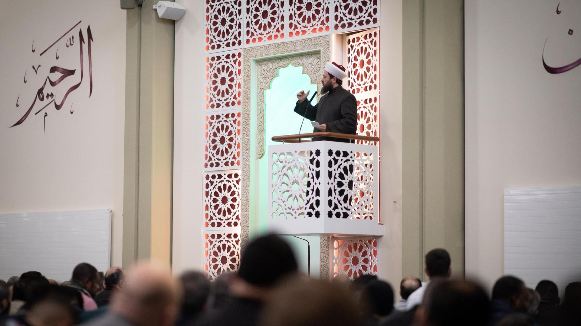 Ein Imam steht auf einem kleinen verzierten Balkon und spricht zu einer Gruppe Männer. An der Wand befinden sich arabische Schriftzeichen. Ein Imam steht auf einem kleinen verzierten Balkon und spricht zu einer Gruppe Männer. An der Wand befinden sich arabische Schriftzeichen.