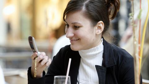Eine junge Frau tippt auf ihrem Mobiltelefon eine SMS, während sie in einem Cafe sitzt.