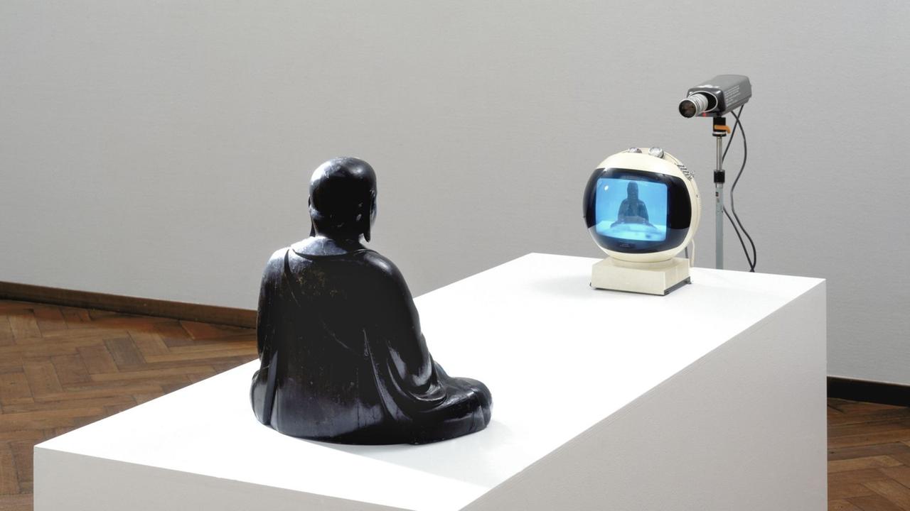Die Installation TV-Buddha (1974) von Nam June Paik. - Eine Buddhafigur scheint sich selbst in einem Fernseher zu betrachten. Die Installation TV-Buddha (1974) von Nam June Paik. - Eine Buddhafigur scheint sich selbst in einem Fernseher zu betrachten.