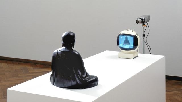 Die Installation TV-Buddha (1974) von Nam June Paik. - Eine Buddhafigur scheint sich selbst in einem Fernseher zu betrachten.