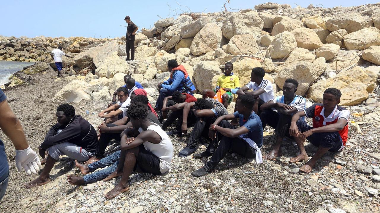 Eine Gruppe von Flüchtlingen sitzt nach einem Schiffsunglück auf Steinen an einem Strand in Libyen. Eine Gruppe von Flüchtlingen sitzt nach einem Schiffsunglück auf Steinen an einem Strand in Libyen.