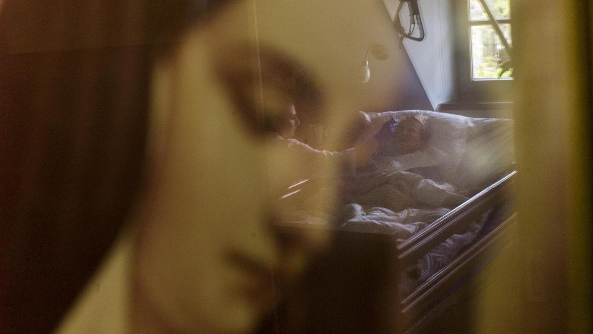 Auf dem Foto hinter dem gemalten Bildnis einer Frau in der Unschärfe eine ältere Dame mit einer Pfegerin am Sterbebett in häuslicher Umgebung zu sehen. Auf dem Foto hinter dem gemalten Bildnis einer Frau in der Unschärfe eine ältere Dame mit einer Pfegerin am Sterbebett in häuslicher Umgebung zu sehen.
