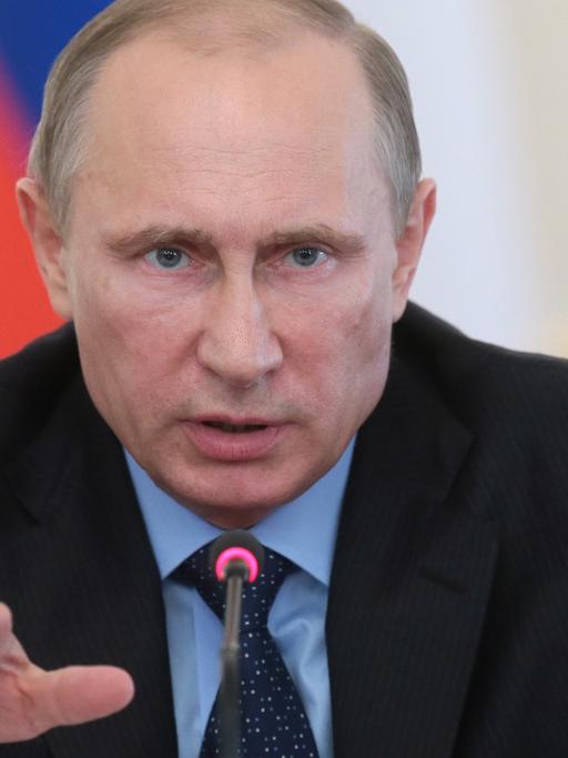 Wladimir Putin sitzt an einem Mikrofon, gestikuliert und spricht