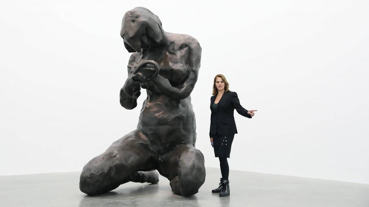 Ein weißer Raum, darin eine sehr große Skulptur, die mit groben Zügen eine menschliche Figur markiert, daneben etwa halb so groß die Künstlerin Tracey Emin. Ein weißer Raum, darin eine sehr große Skulptur, die mit groben Zügen eine menschliche Figur markiert, daneben etwa halb so groß die Künstlerin Tracey Emin.