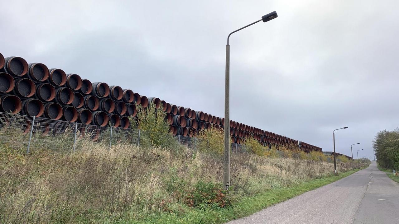 Große übereinandergestapelte, schwarze Rohre lagern hinter einem Metallzaum. Rechts daneben ist ein Gehweg mit Straßenlaternen zu sehen. Große übereinandergestapelte, schwarze Rohre lagern hinter einem Metallzaum. Rechts daneben ist ein Gehweg mit Straßenlaternen zu sehen.