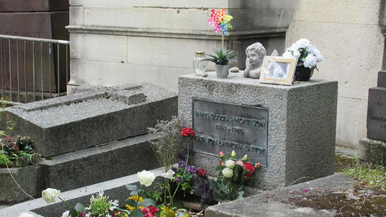 Ein Grab mit einem mittelhohen rechteckigen Grabstein, auf dem ein Foto des Sängers, Ein steinerner Engelskopf und ein buntes Windrad stehen. Auf dem Grab wachsen einige weiße und rote Rosen.